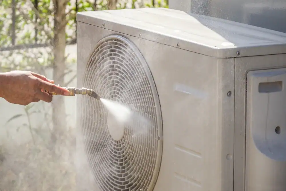 A person sprays water to clean the outdoor unit of an air conditioner, focusing on the fan and grille area.
