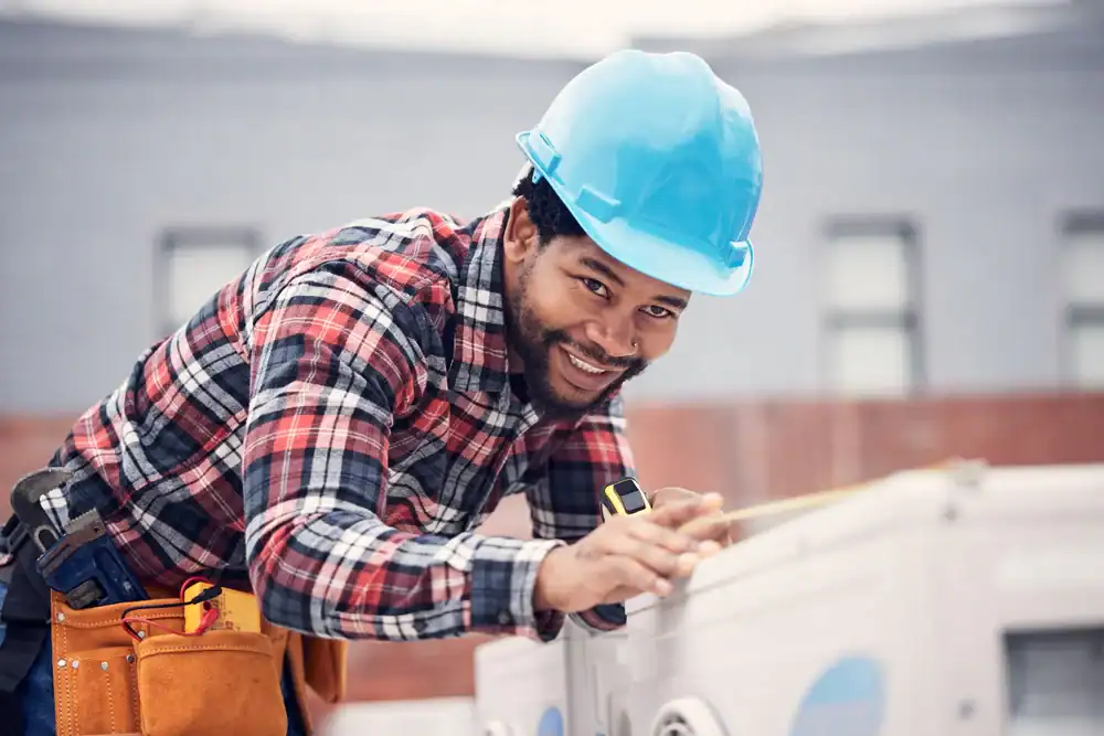 A smiling construction worker in a blue hard hat and plaid shirt uses a measuring tape while working outdoors. He has a tool belt on and is leaning over a surface, focused on his task.
