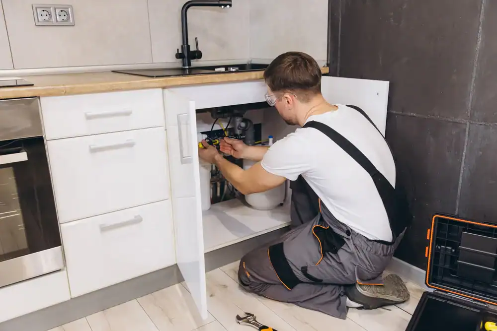 A plumber wearing overalls and safety glasses kneels on the floor, working under a kitchen sink with tools. An open toolbox and wrench are nearby.