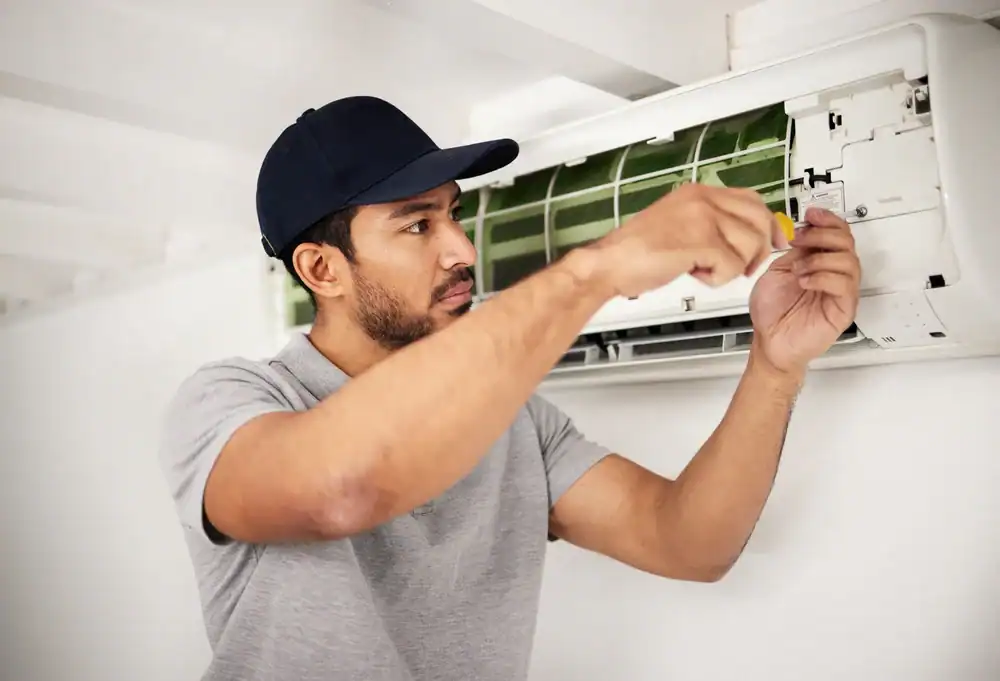 A man wearing a dark cap and gray polo shirt repairs an air conditioning unit mounted on a wall, using a screwdriver and focusing on the device's internal components.