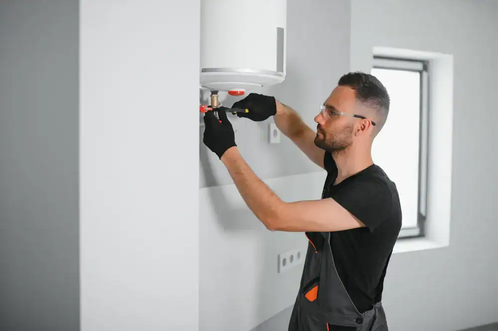 A man wearing safety glasses and gloves installs or repairs a water heater mounted on a wall inside a modern, bright room.
