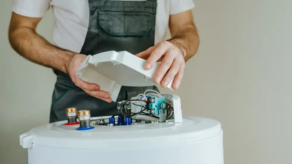 A person in work overalls opens the top cover of a water heater, exposing internal electrical components and connections.